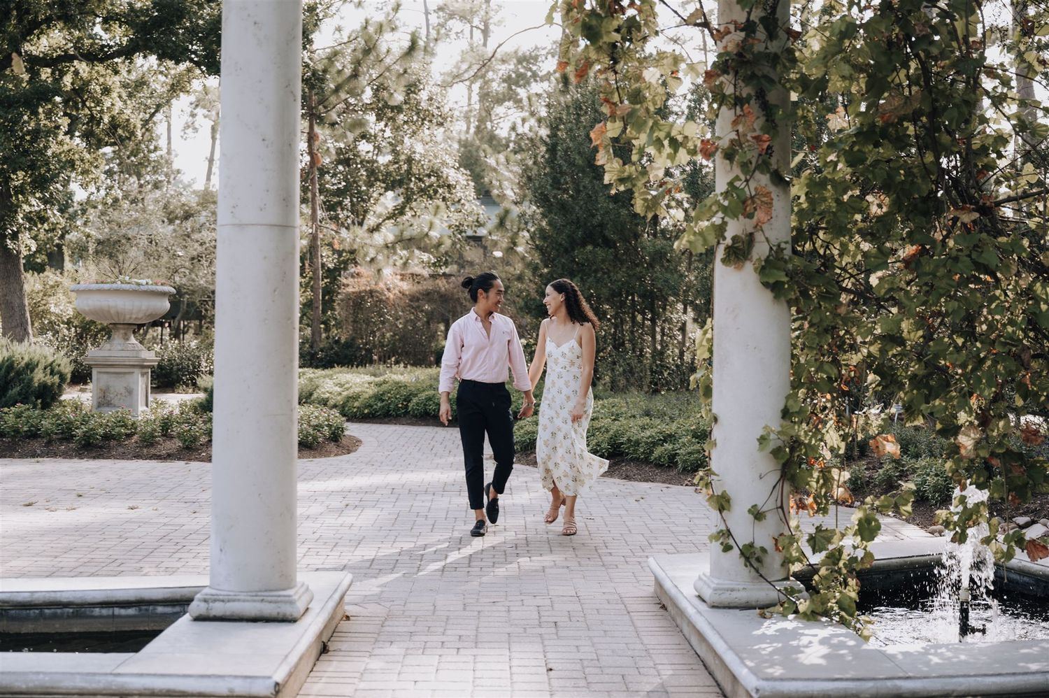 A couple strolls together along a scenic garden path lined with white pillars and lush greenery in an elegant outdoor setting.