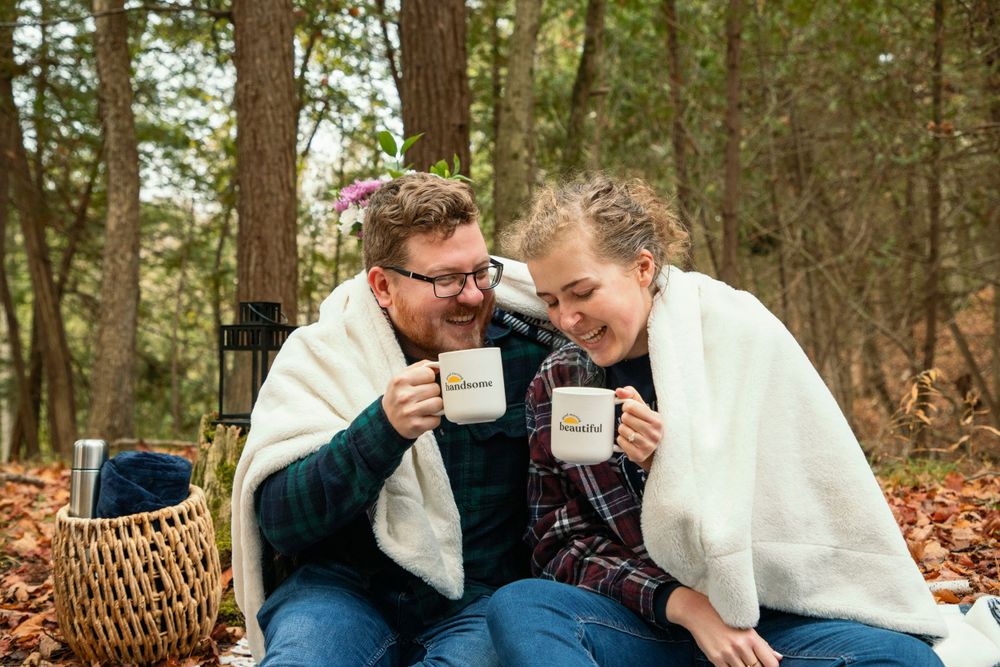 in a forested area an engaged couple sitting next to each other wrapped with a white blanket, and drinking  hot chocolate
