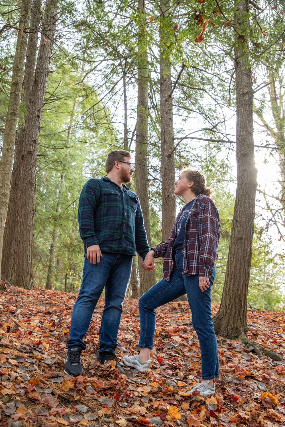 A couple in a forrest of red leaves and tall mostly bare trees (some evergreens) standing next to one another on a hill gazing at each other