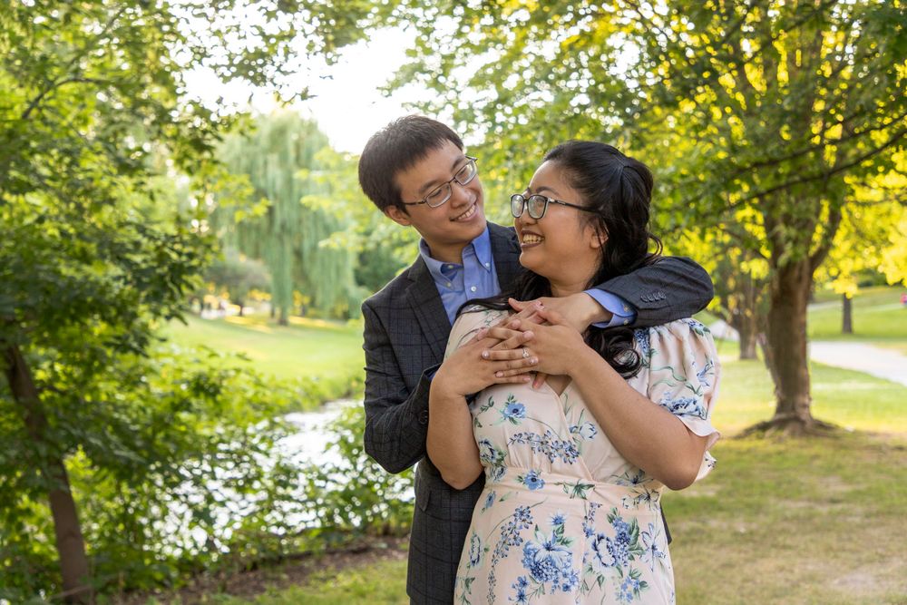 An image of a couple with the guy behind her wrapping his arms around her neck and shoulders looking at one another