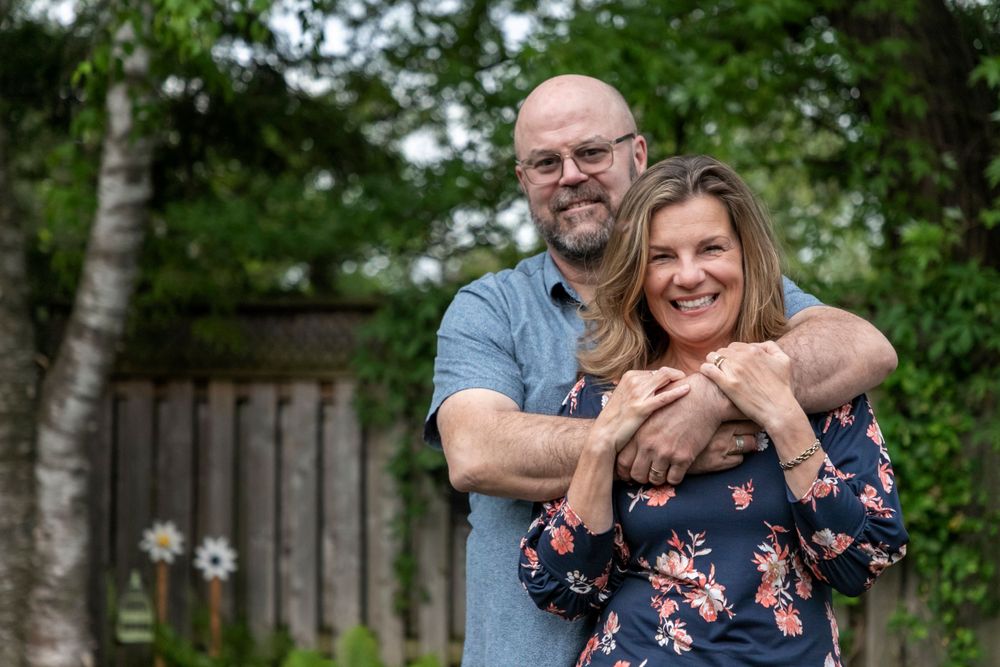 a photograph of a man with his lady standing in front of him; he's wrapping his arms around his shoulders and she is holding his wrists