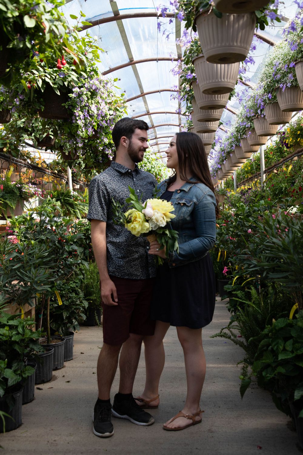 a young couple in the aisle of a greenhouse holding flowers