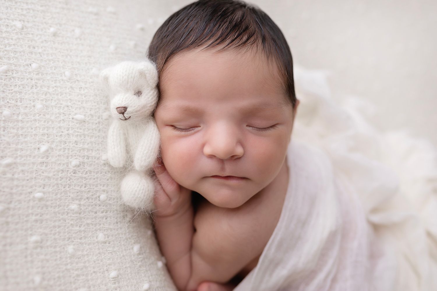 Sleeping newborn with tiny white teddy bear companion wrapped in soft white blanket.