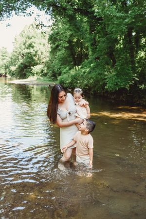 Family playing and splashing in shallow creek water surrounded by green trees.