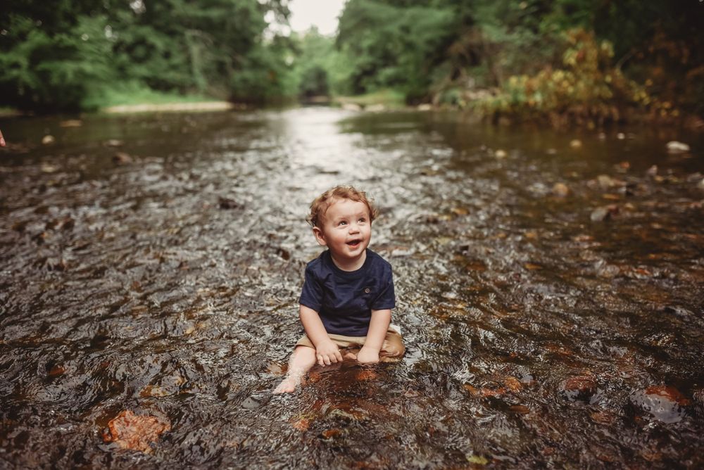 Child playing in a shallow stream with rocks and trees in the background.