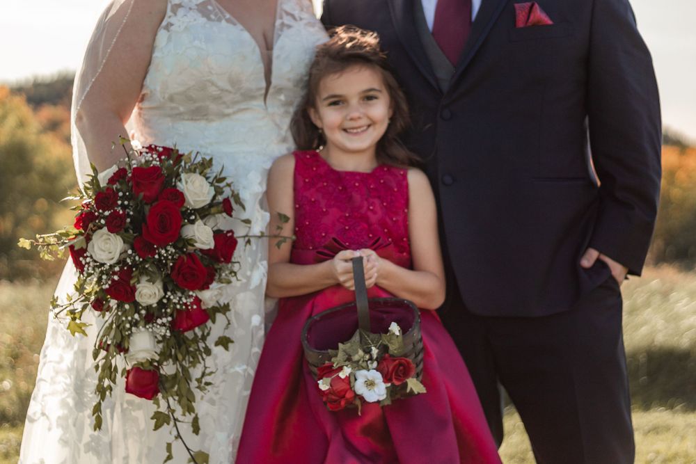 Wedding couple and flower girl in red dress posing with bridal bouquet of red and white flowers.