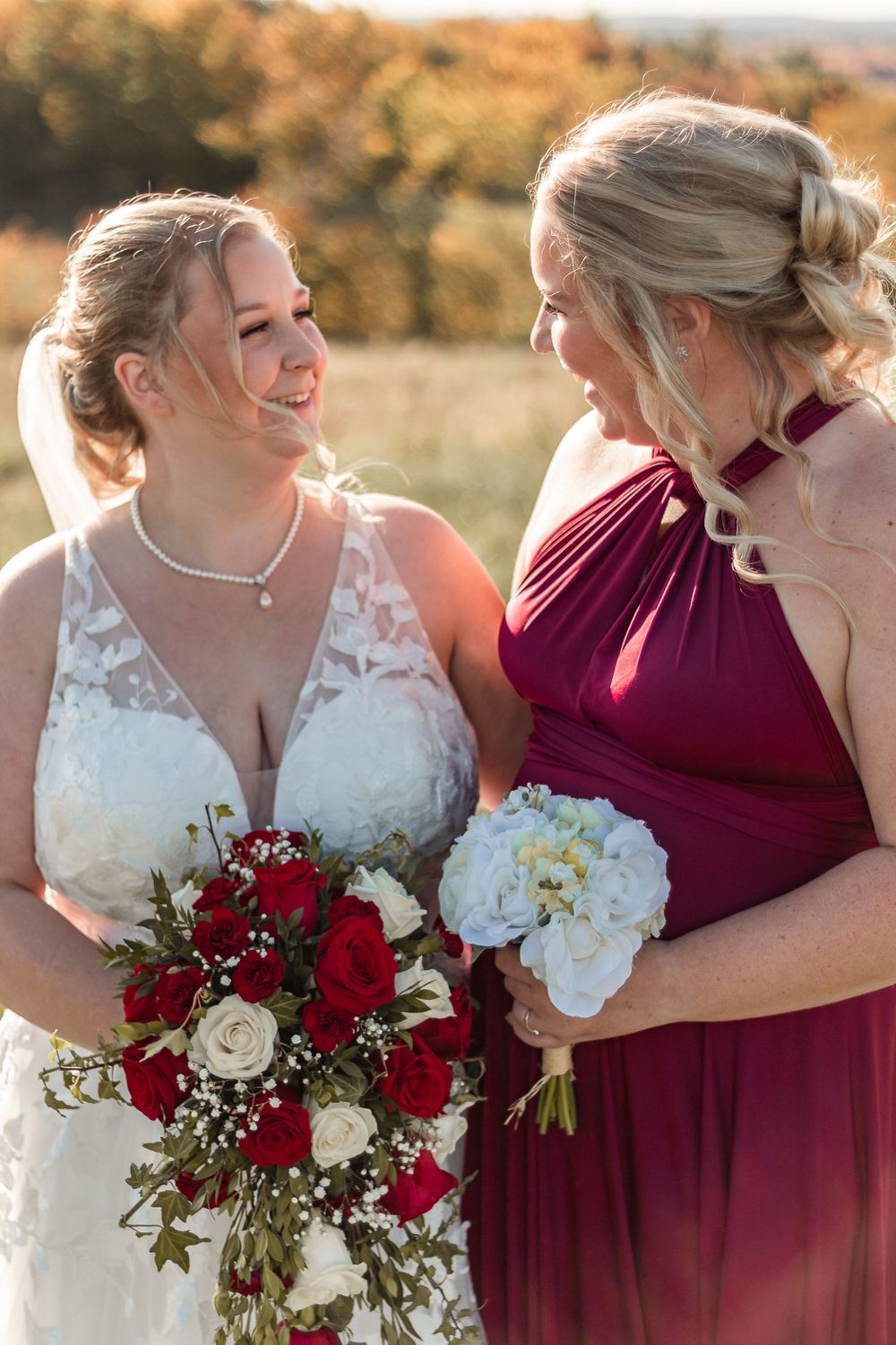 Bride and bridesmaid sharing a joyful moment with red and white bouquets in autumn setting.