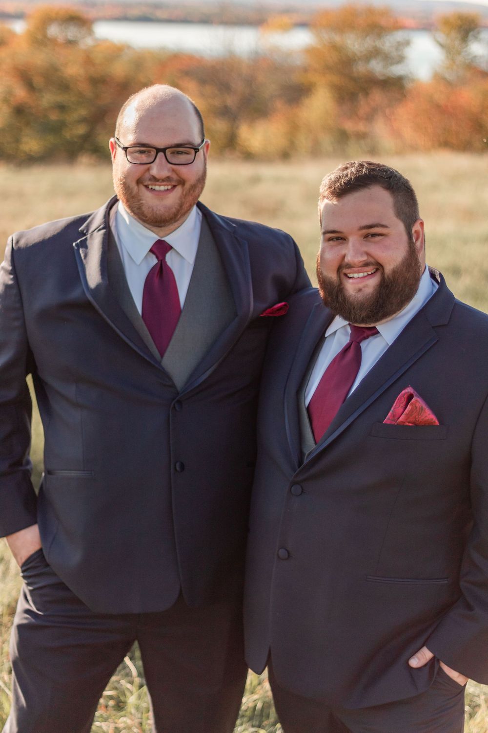 Two groomsmen in dark suits and burgundy ties smile together in autumn setting.