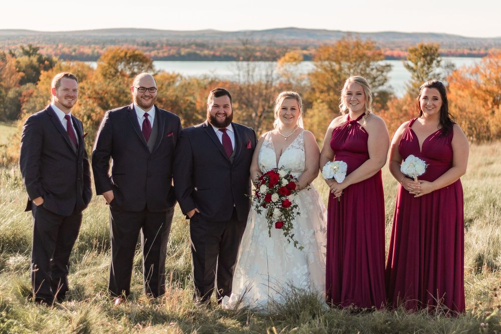 Wedding party in burgundy and black attire pose together against fall landscape.