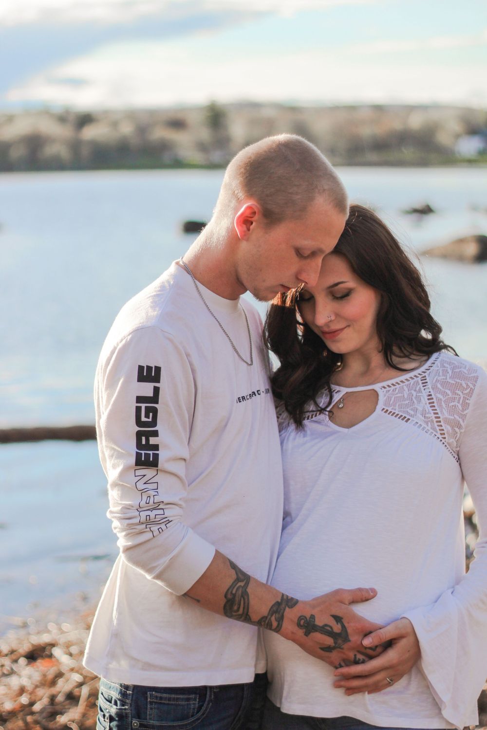 A tender maternity photo of a couple in white clothing by the waterfront.