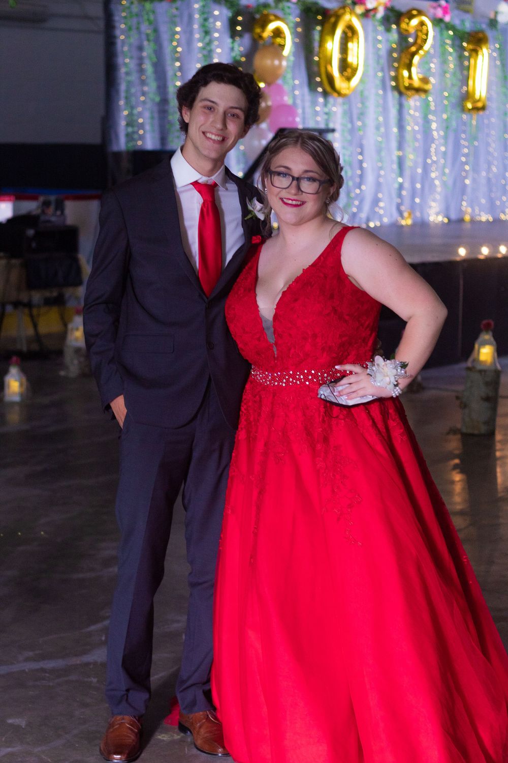 Formal couple poses at event with gold balloon decorations and sparkly backdrop.