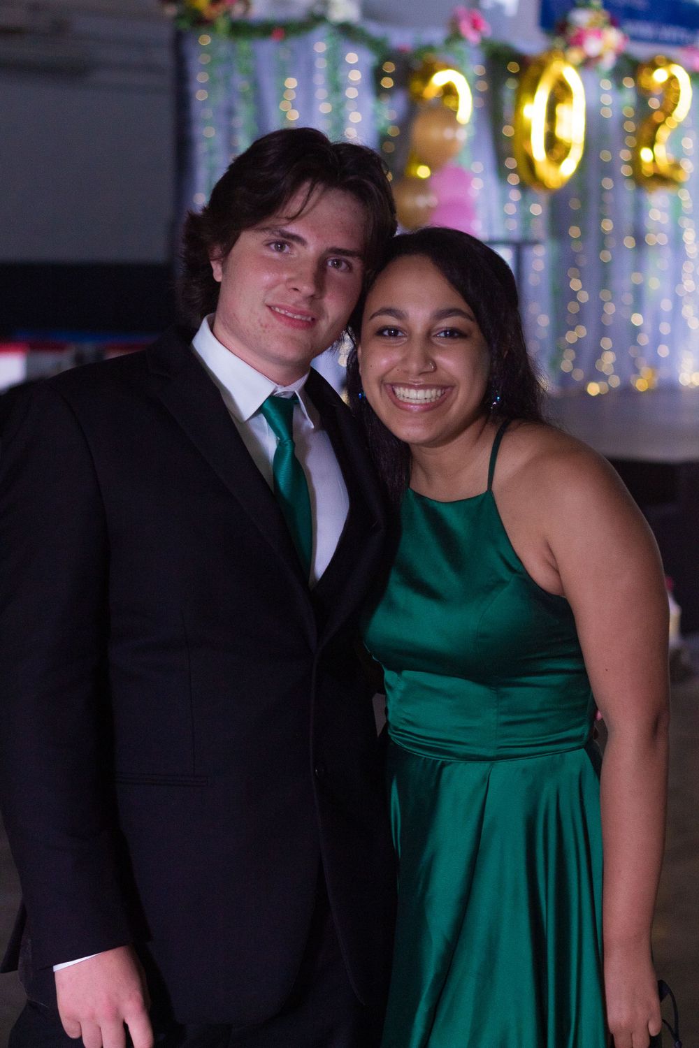 Couple in formal attire poses at event with gold decorations in background.