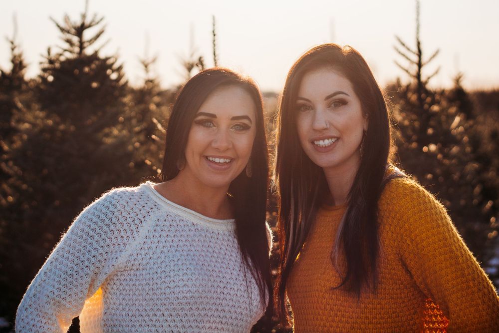 Two friends pose together outdoors with evergreen trees in background.