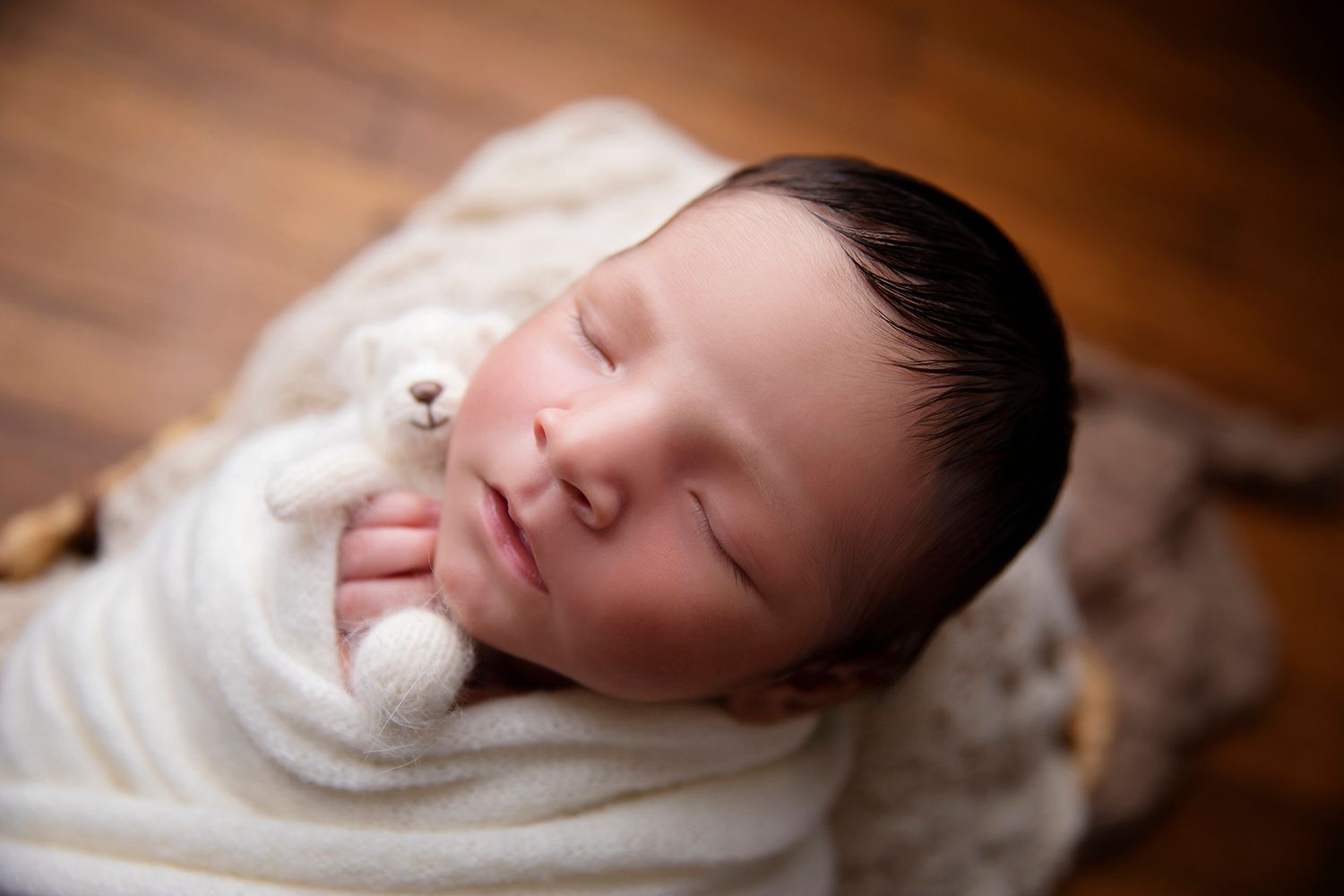 Newborn wrapped in white holding tiny teddy bear on wooden backdrop.