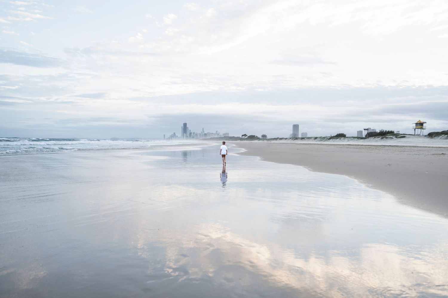 Solitary figure walking on misty beach with city skyline in distant background.