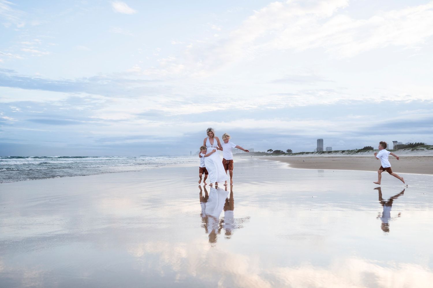 Beach family portrait with mirror-like water reflection against cloudy coastal sky.