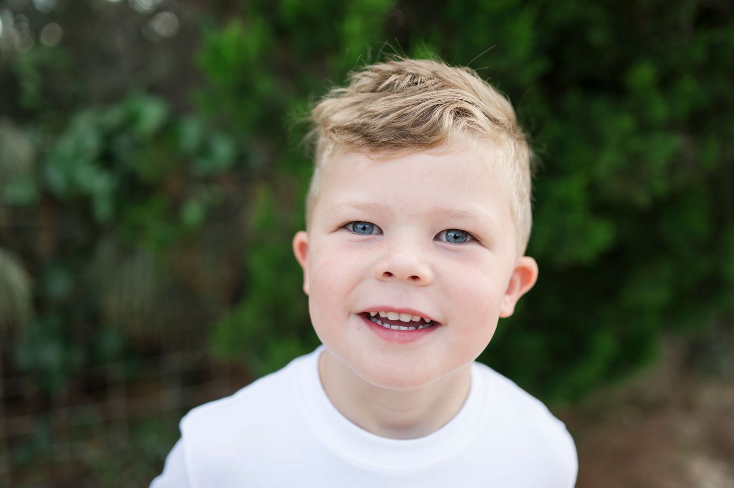 Young child in white shirt smiling outdoors with green foliage background.