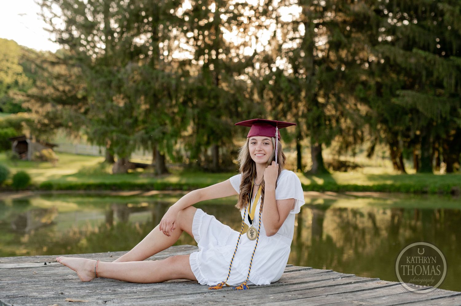 Graduate in a white dress relaxes on a wooden dock by a serene pond during a sunset photo session.