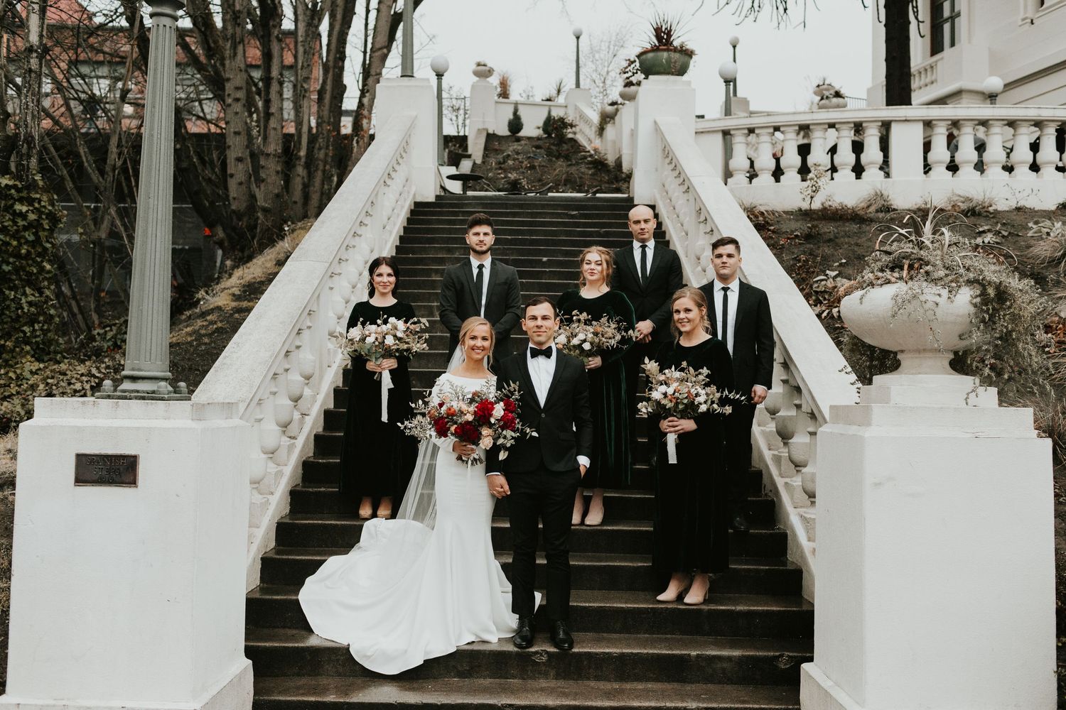 Wedding party gathered on white stone staircase for formal group photo.