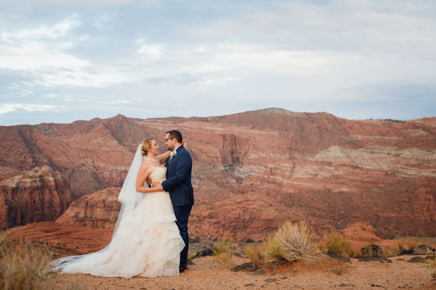 Bride and groom embracing on a scenic overlook with vast red canyon landscape and dramatic cloudy sky backdrop.