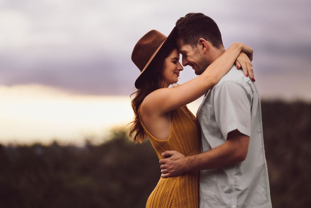 A couple embraces closely, smiling with foreheads touching, as the woman wears a hat and shows her ring.