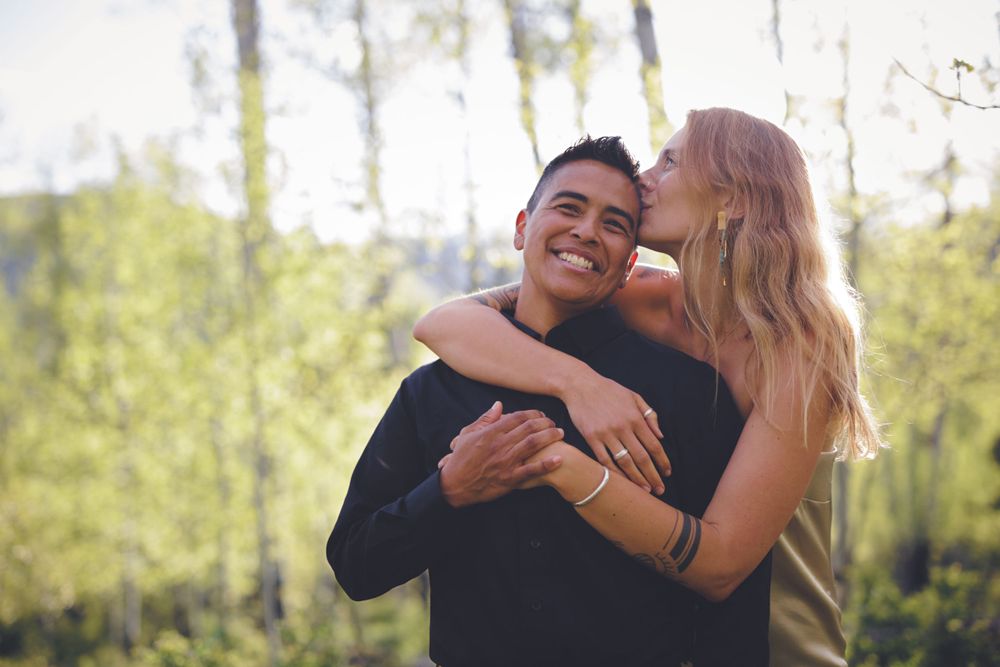 Happy couple embracing outdoors, woman kissing mans cheek while he smiles in a sunny forest setting.
