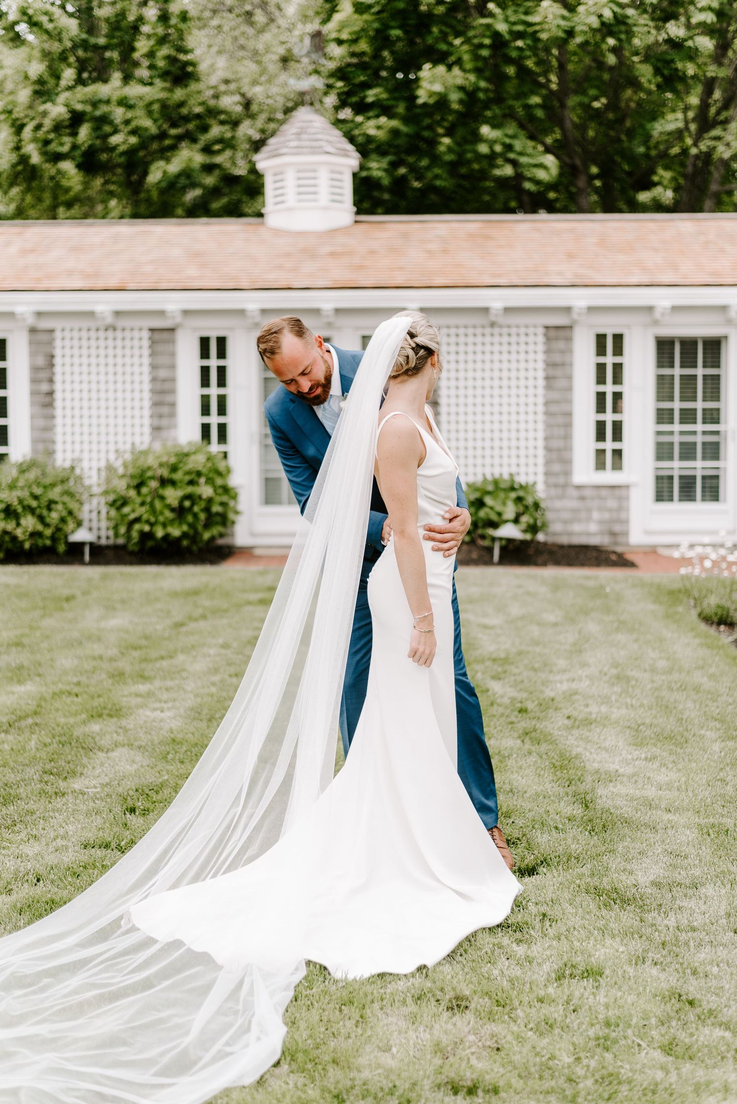 An elegant wedding couple embraces on manicured lawn with flowing cathedral veil and classic architecture.