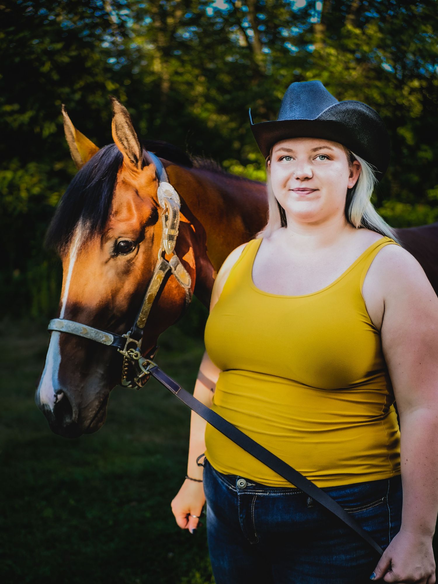 Beautiful chestnut horse with bridle standing next to person in yellow tank top and cowboy hat.