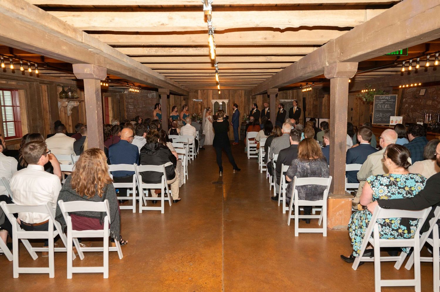 Indoor wedding ceremony in rustic venue with wooden beams and rows of white chairs.