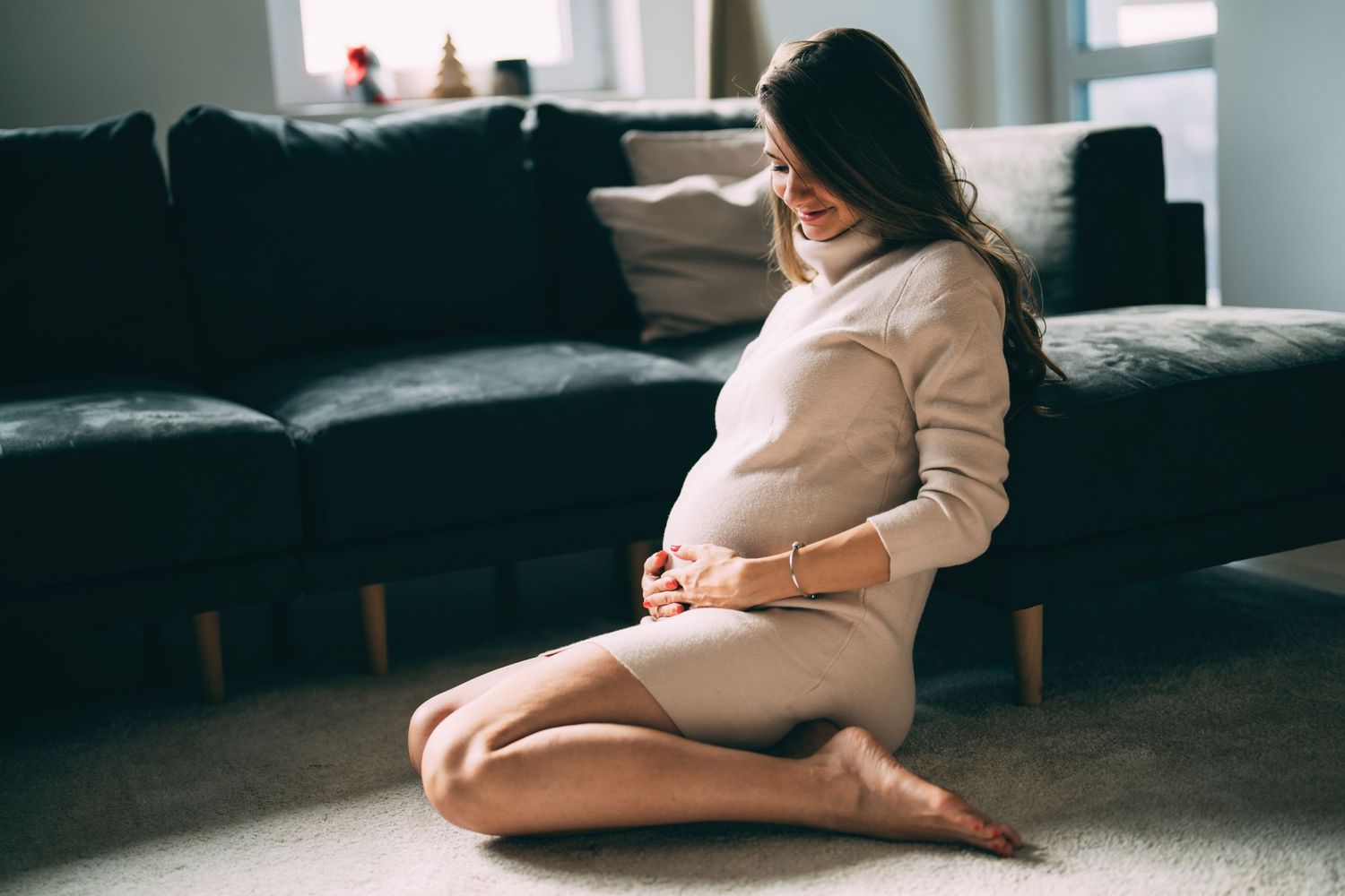 Peaceful maternity scene with pregnant person in beige sweater sitting on dark sofa.