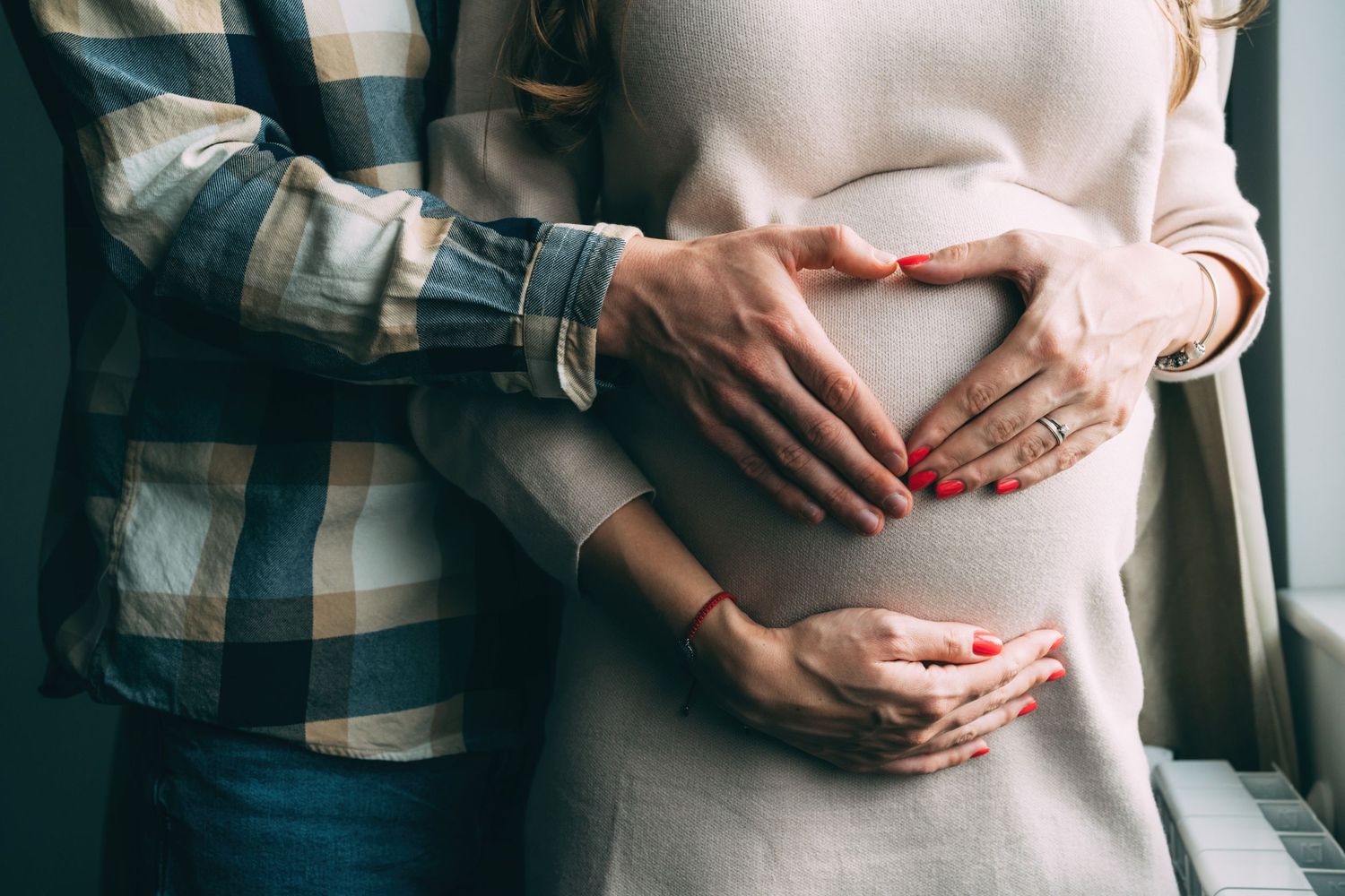 Close-up of hands forming heart shape on pregnant belly against beige sweater.