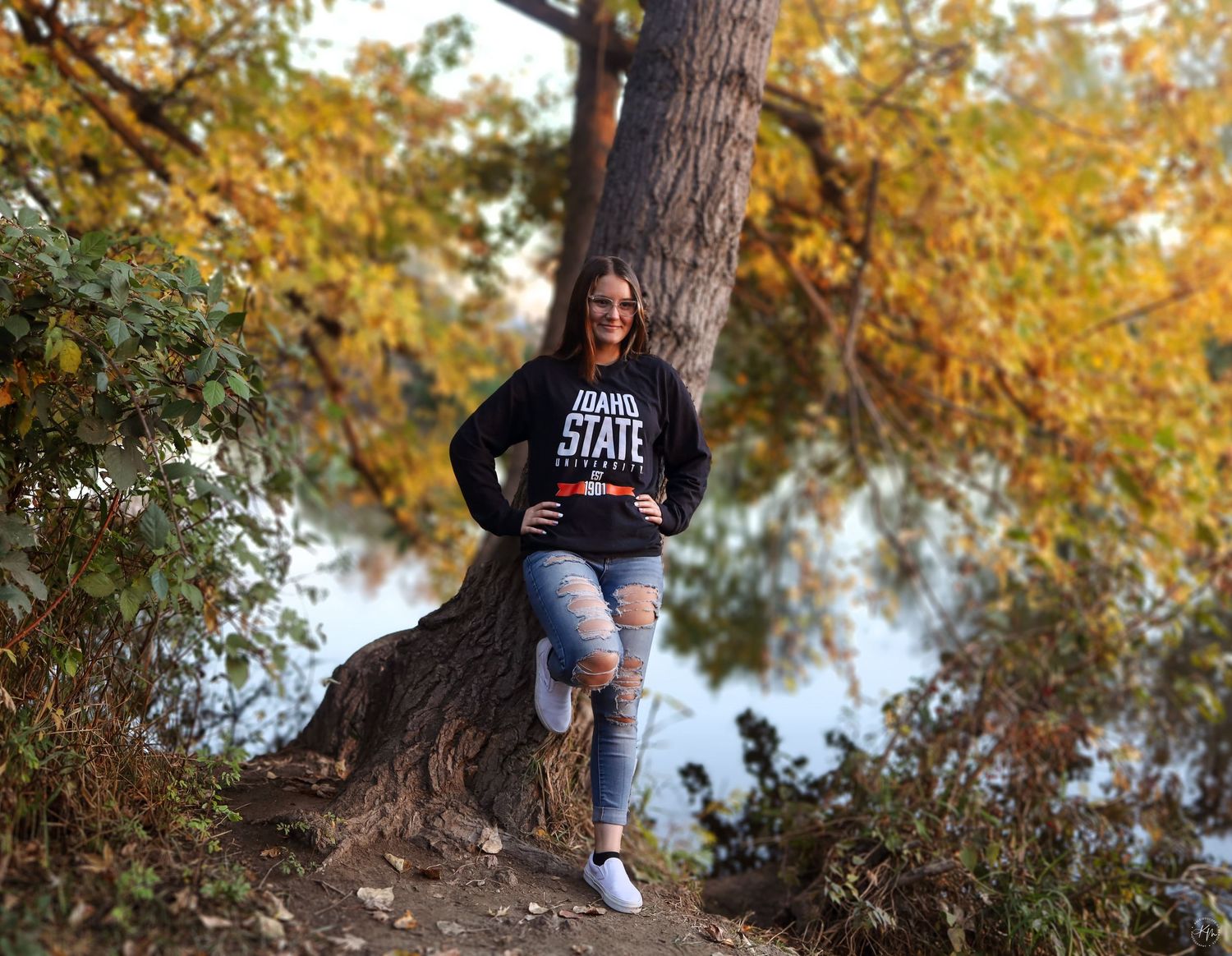 A person wearing a varsity sweatshirt poses casually against a tree trunk near a lake during autumn.