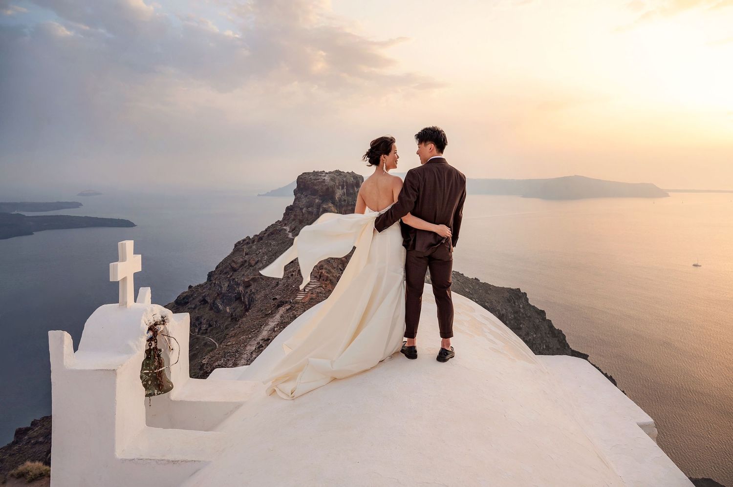 A wedding couple stands atop a white cliffside in Santorini during sunset overlooking the Mediterranean Sea.