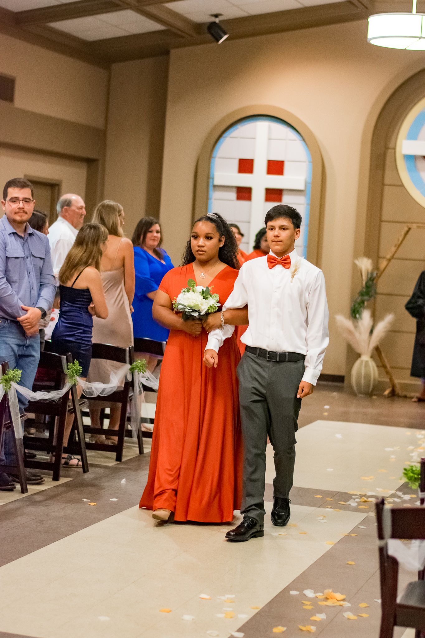 Wedding party members in orange and formal attire walk down the aisle during ceremony.