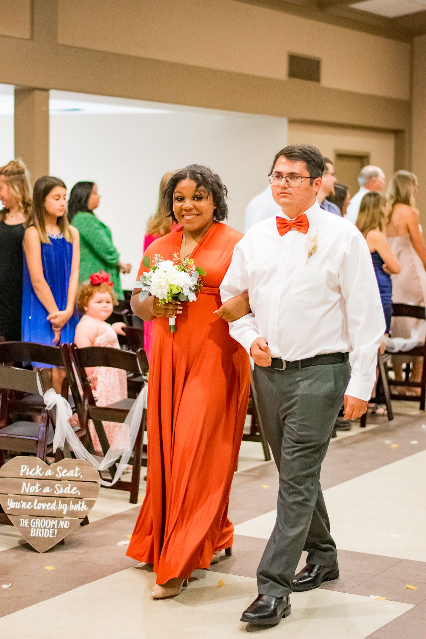 Wedding attendants in orange and white attire process down church aisle carrying bouquets.