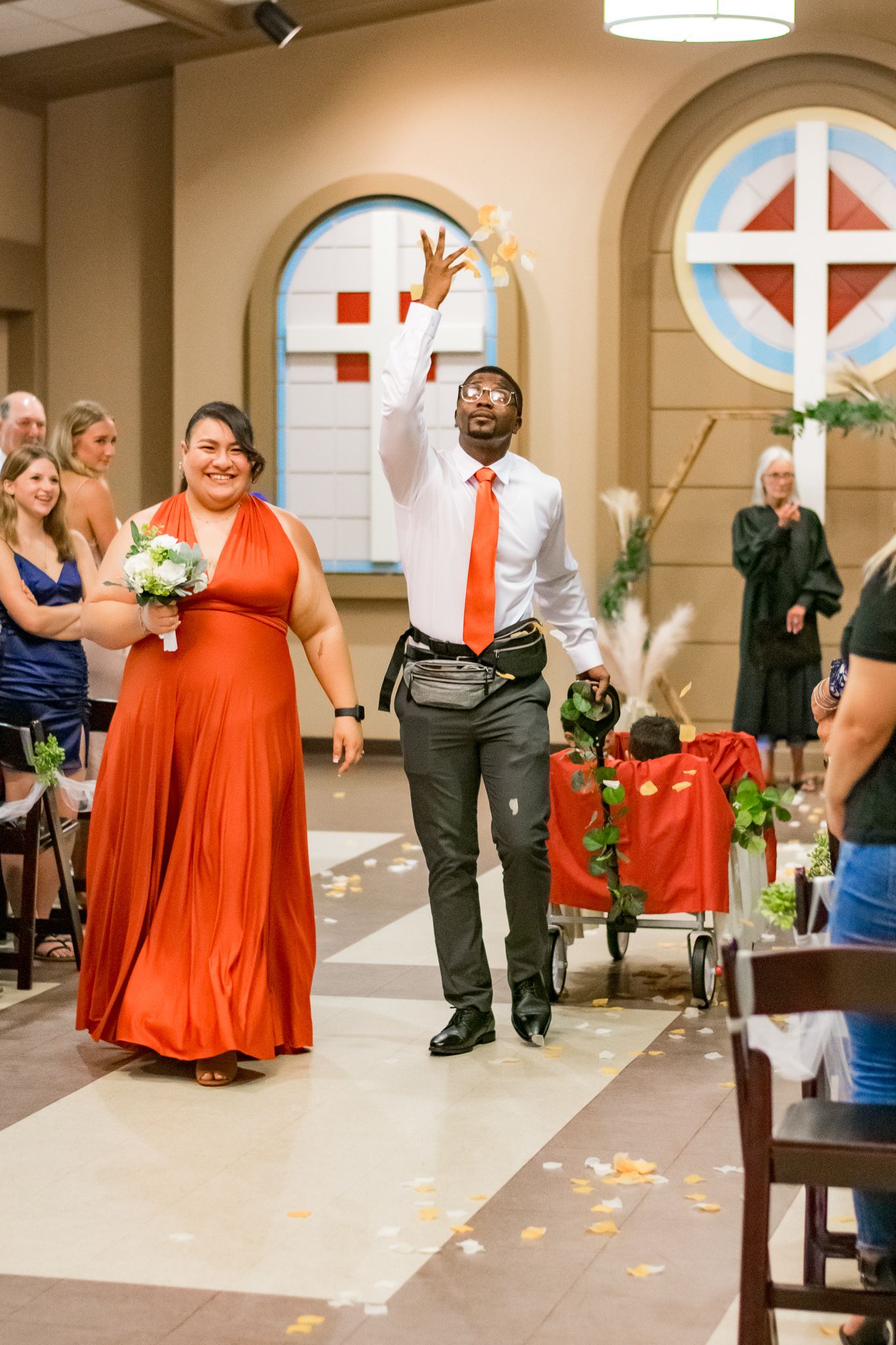Wedding party member in orange dress walks down aisle while another celebrates enthusiastically.