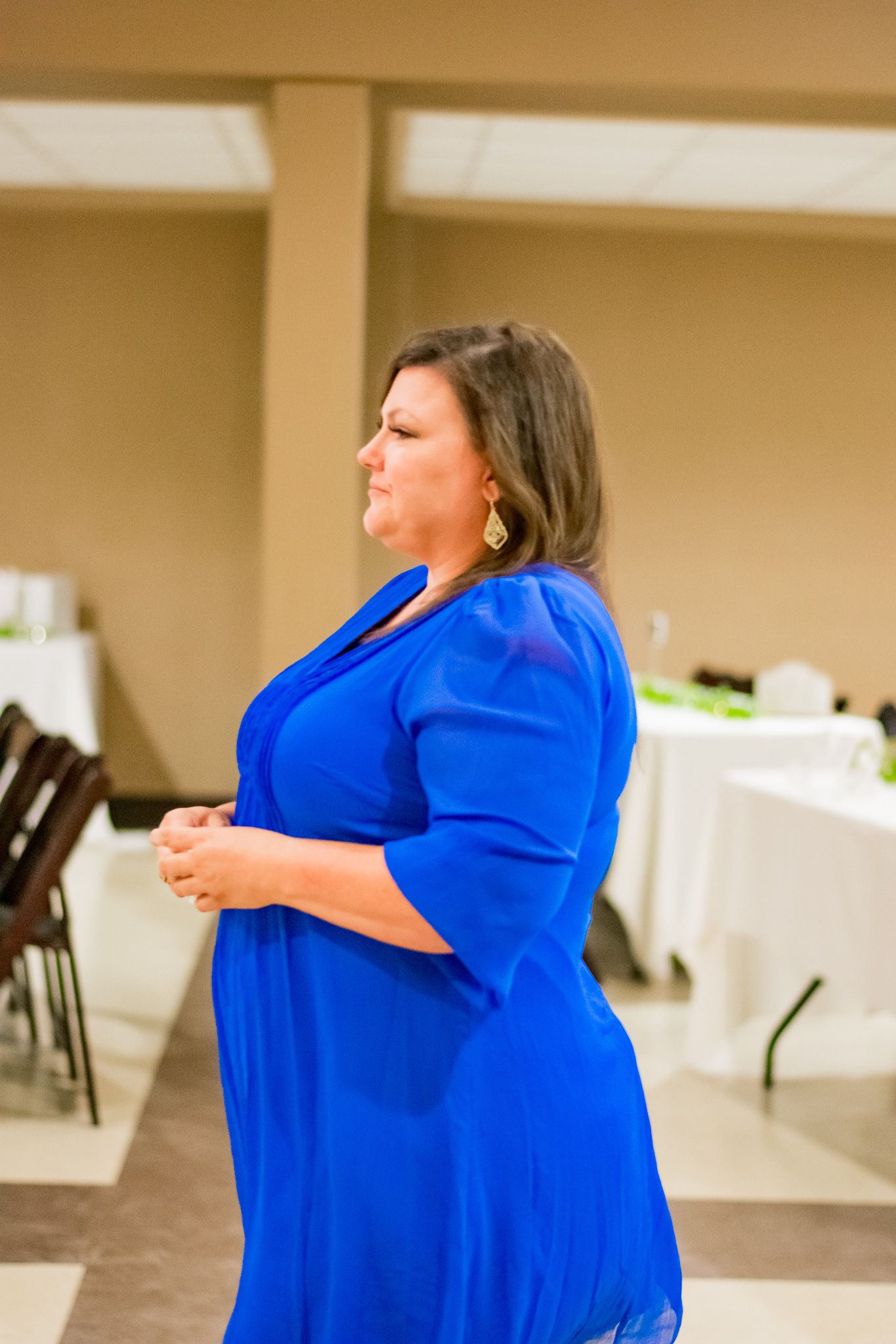 Woman in bright blue dress stands in church reception hall with tables and chairs.
