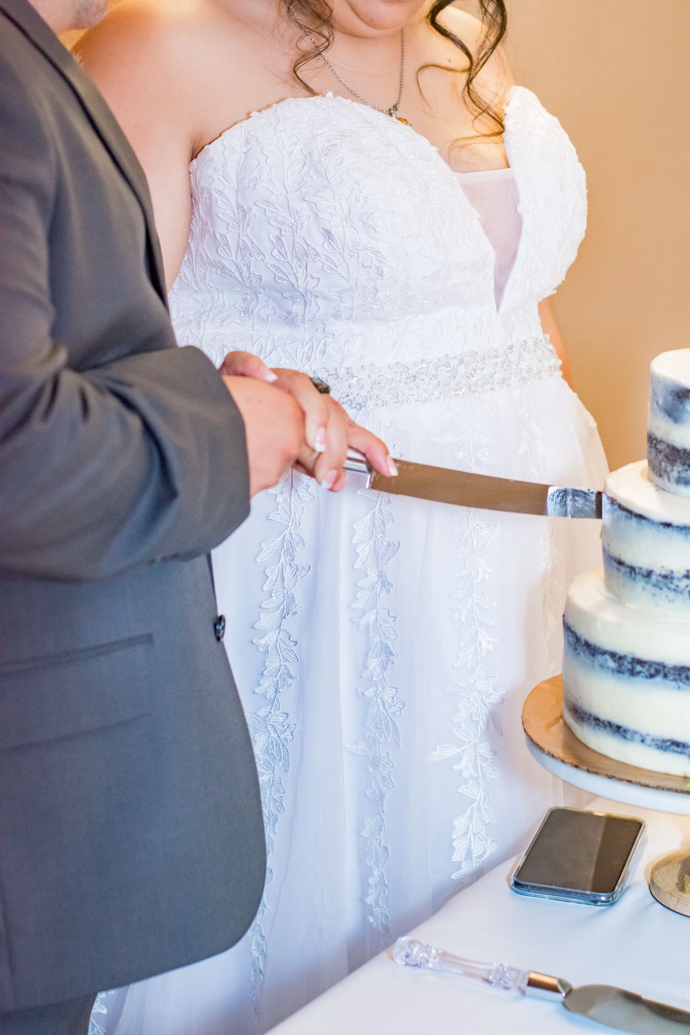 Close-up of a white lace wedding dress and hands cutting into a blue and white tiered wedding cake.