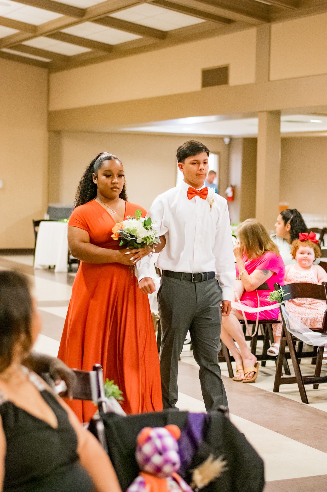 Wedding party members in red dress and bow tie walking down church aisle during ceremony.