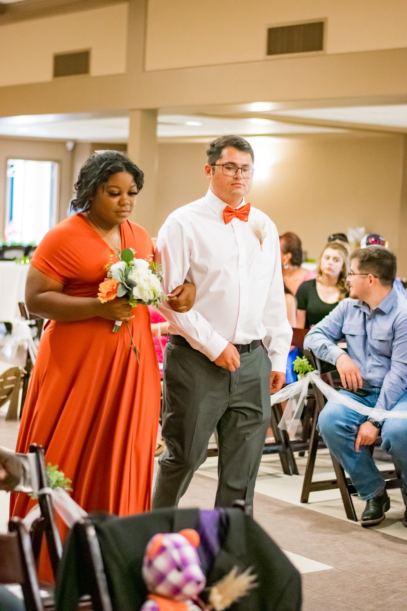 Bridesmaid in flowing red dress and groomsman in bow tie walking down aisle during wedding ceremony.