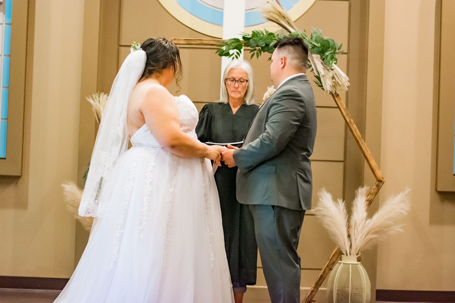 Wedding ceremony taking place under decorative arch with pampas grass arrangements.