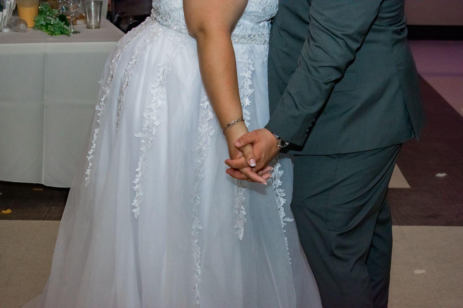 Close-up of joined hands in formal wedding attire against white dress detail.