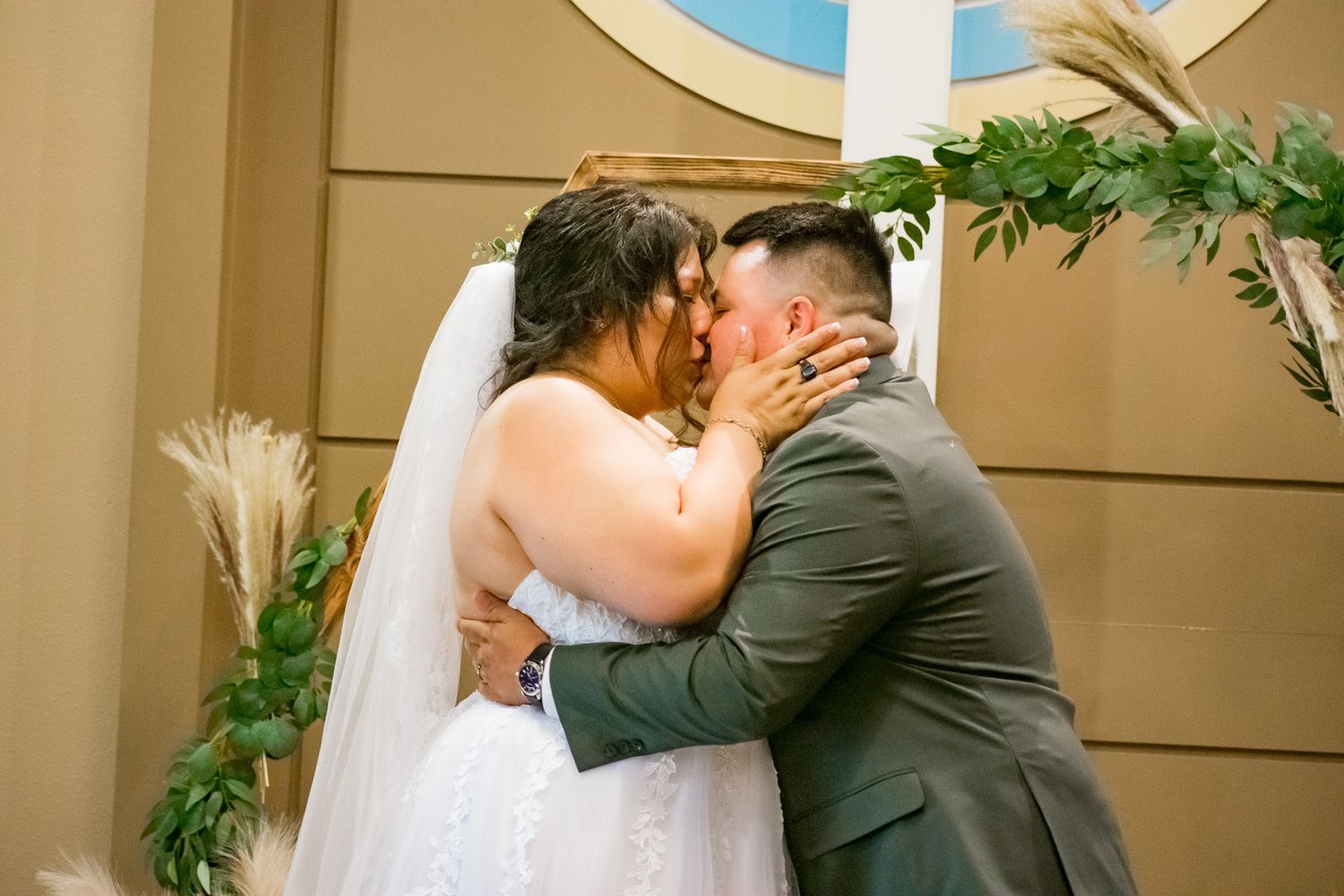 Newlyweds share their first kiss under a decorated wedding arch with greenery and a circular window.