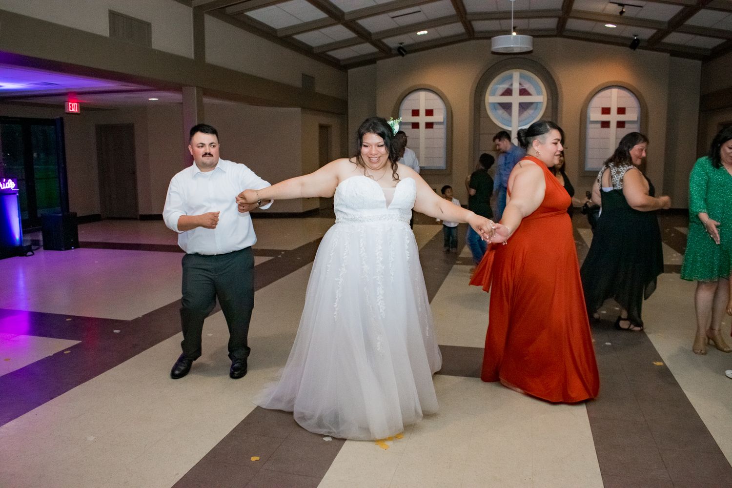 Wedding party dancing together in a church reception hall with purple lighting.