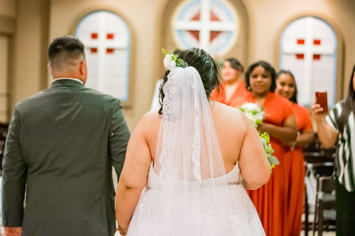 Wedding ceremony in a church with stained glass windows and guests in coral dresses.