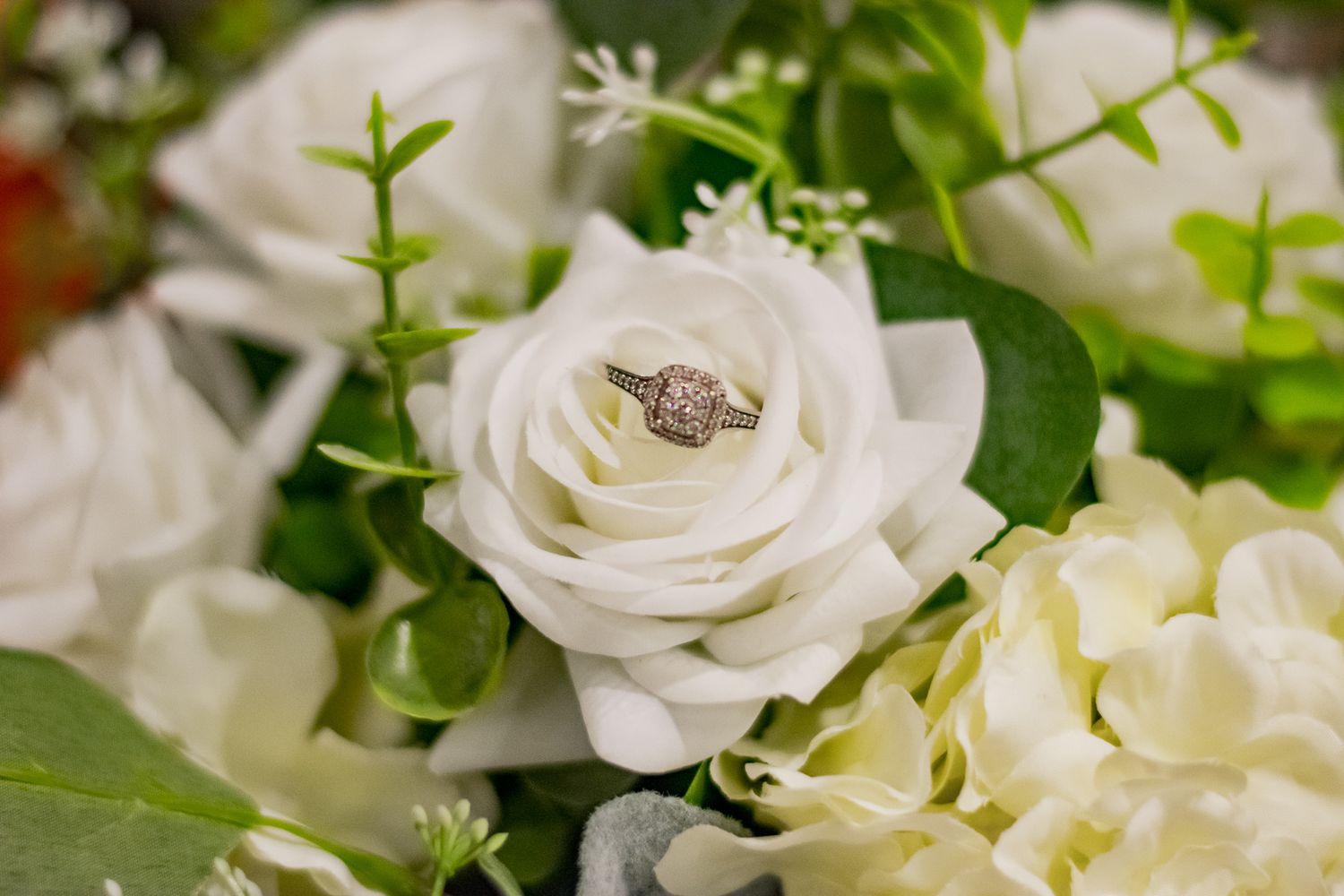 Wedding ring nestled in a white rose surrounded by greenery and floral arrangements.