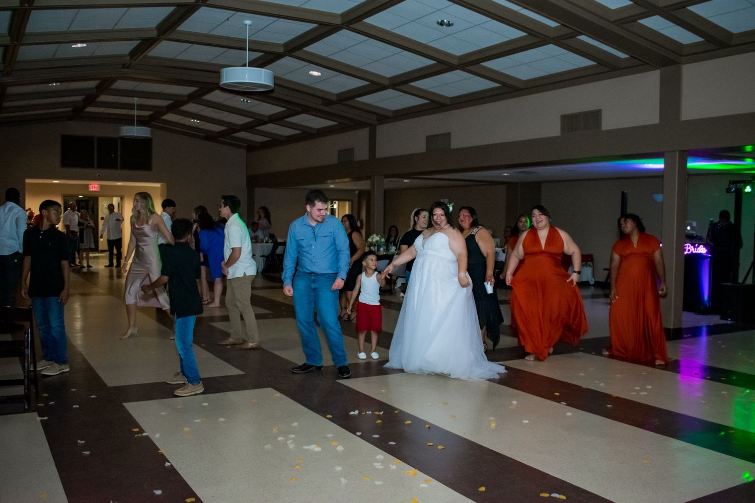 Wedding party dancing in a line on a dance floor with colorful lights in the background.