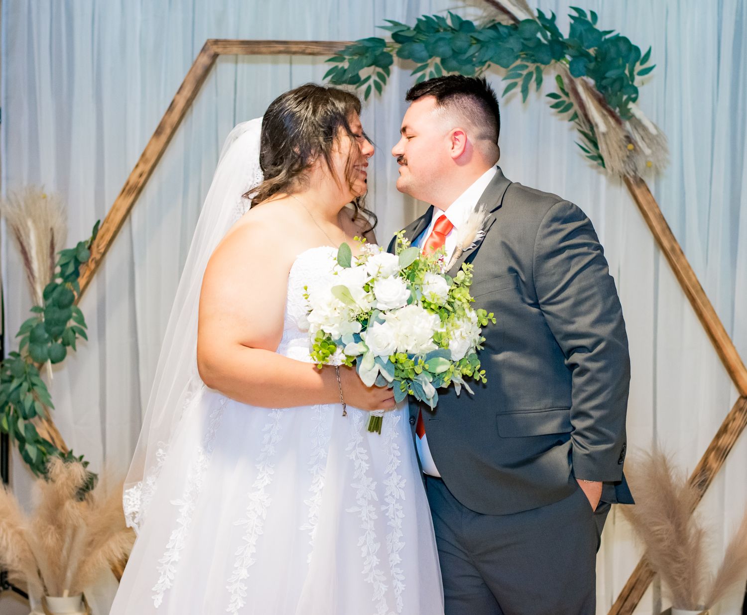 Wedding couple poses under hexagonal arch decorated with greenery and pampas grass.