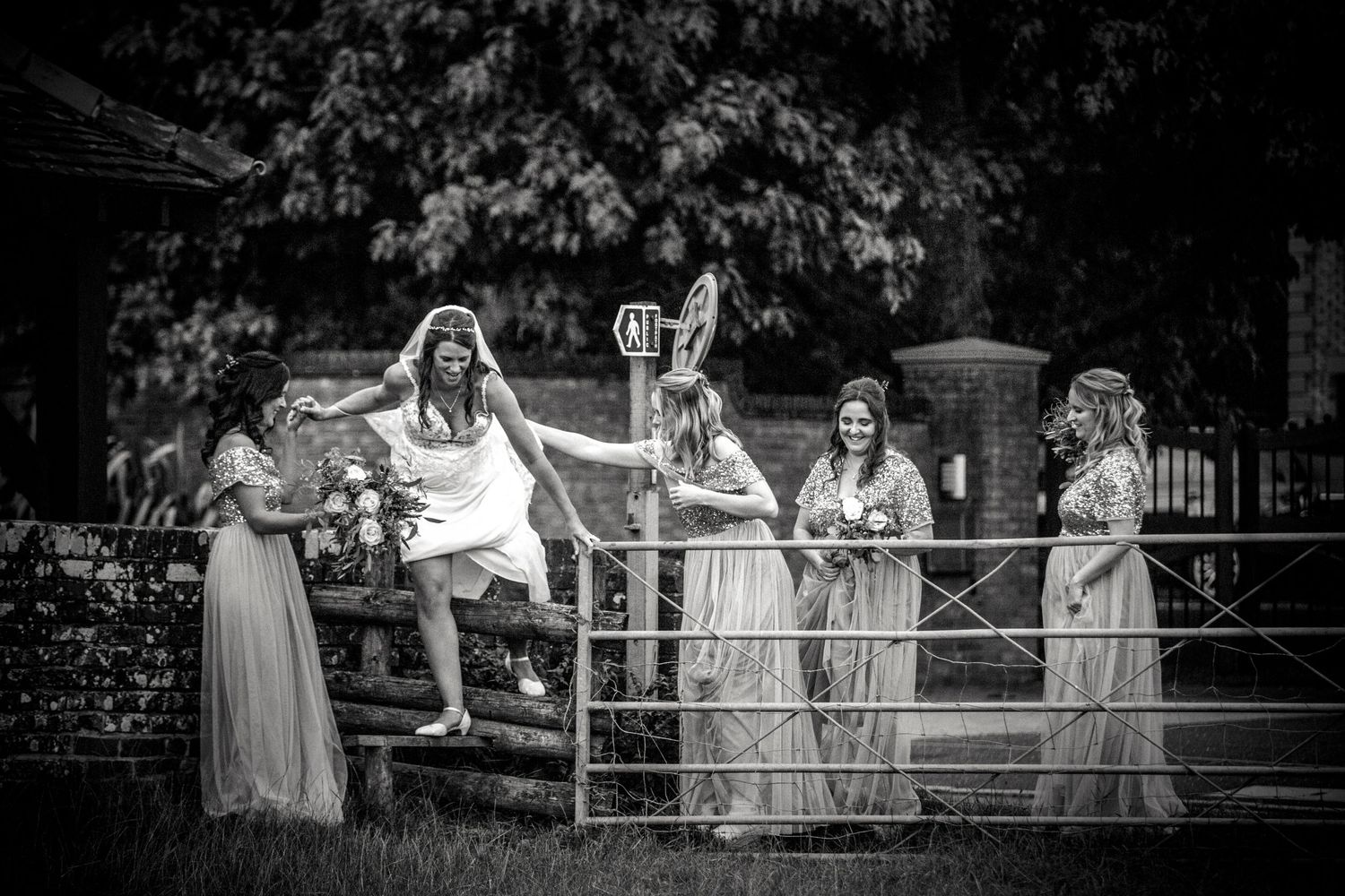 Bridesmaids in flowing dresses pose playfully on a metal gate in black and white photography.
