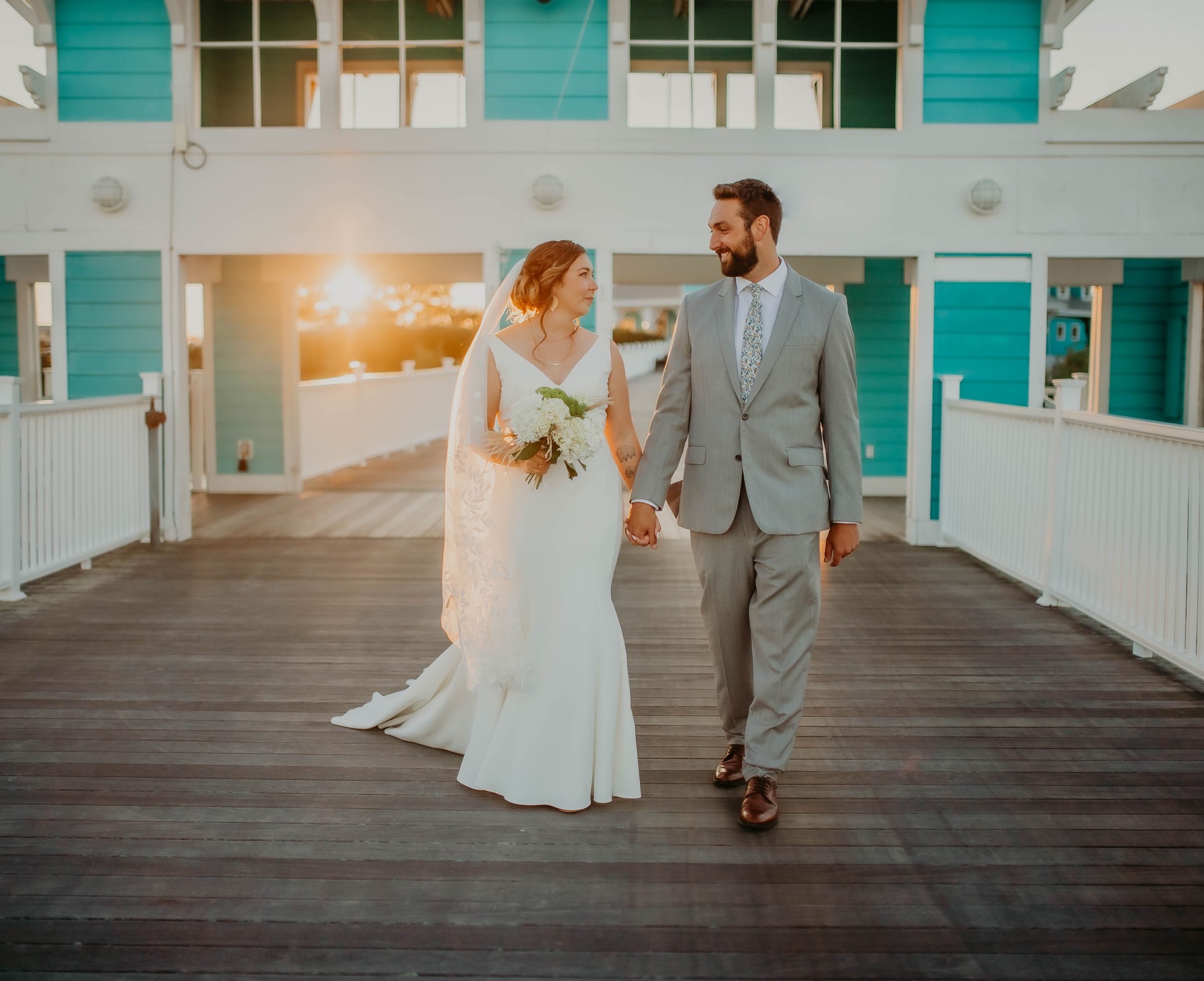 Bride and groom walking hand in hand on a pier at sunset near turquoise coastal buildings.